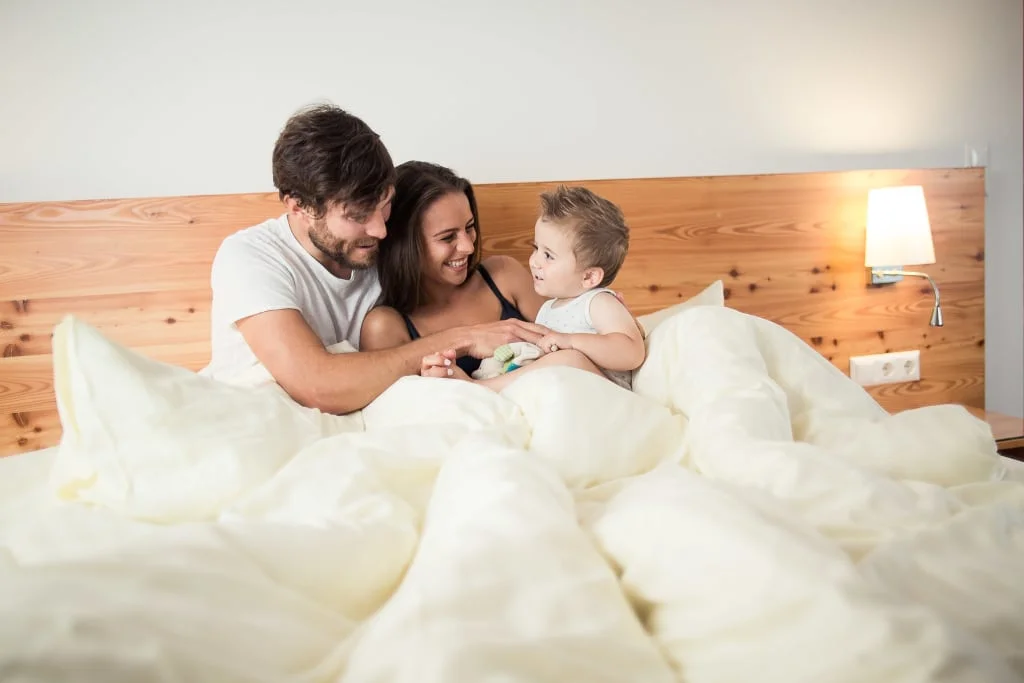 Ouders en kind lachen samen in bed onder een wit dekbed