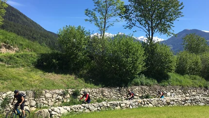 Four cyclists biking on a path beside a stone wall in the mountains
