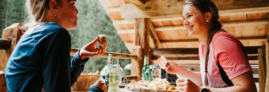 Spätsommer- & Herbstbiken in Toblach © Desire Lines - Tobias Dörr Zwei Frauen essen gemütlich Brot und Käse auf einer Holzterrasse in den Bergen.