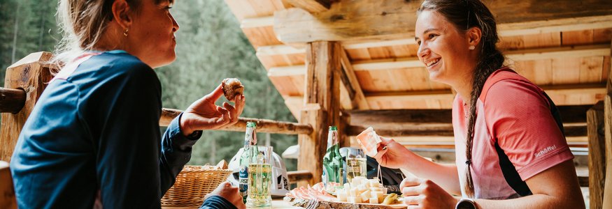 Zwei Frauen essen gemütlich Brot und Käse auf einer Holzterrasse in den Bergen.