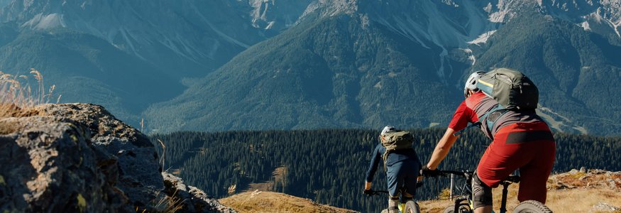 Zwei Mountainbiker fahren auf einem Bergweg mit Alpen im Hintergrund