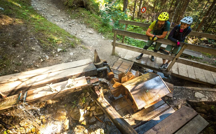 Two cyclists with helmets watching a working water wheel in the forest