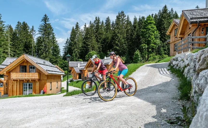 Zwei Radfahrerinnen fahren auf einem Schotterweg vor Holzhäusern und Wald