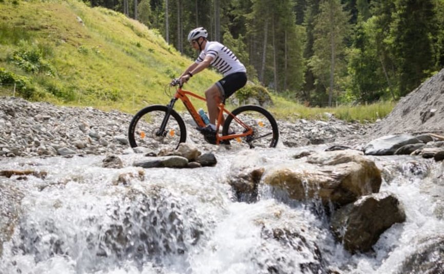 Man riding a mountain bike through a stream in a forested mountain area