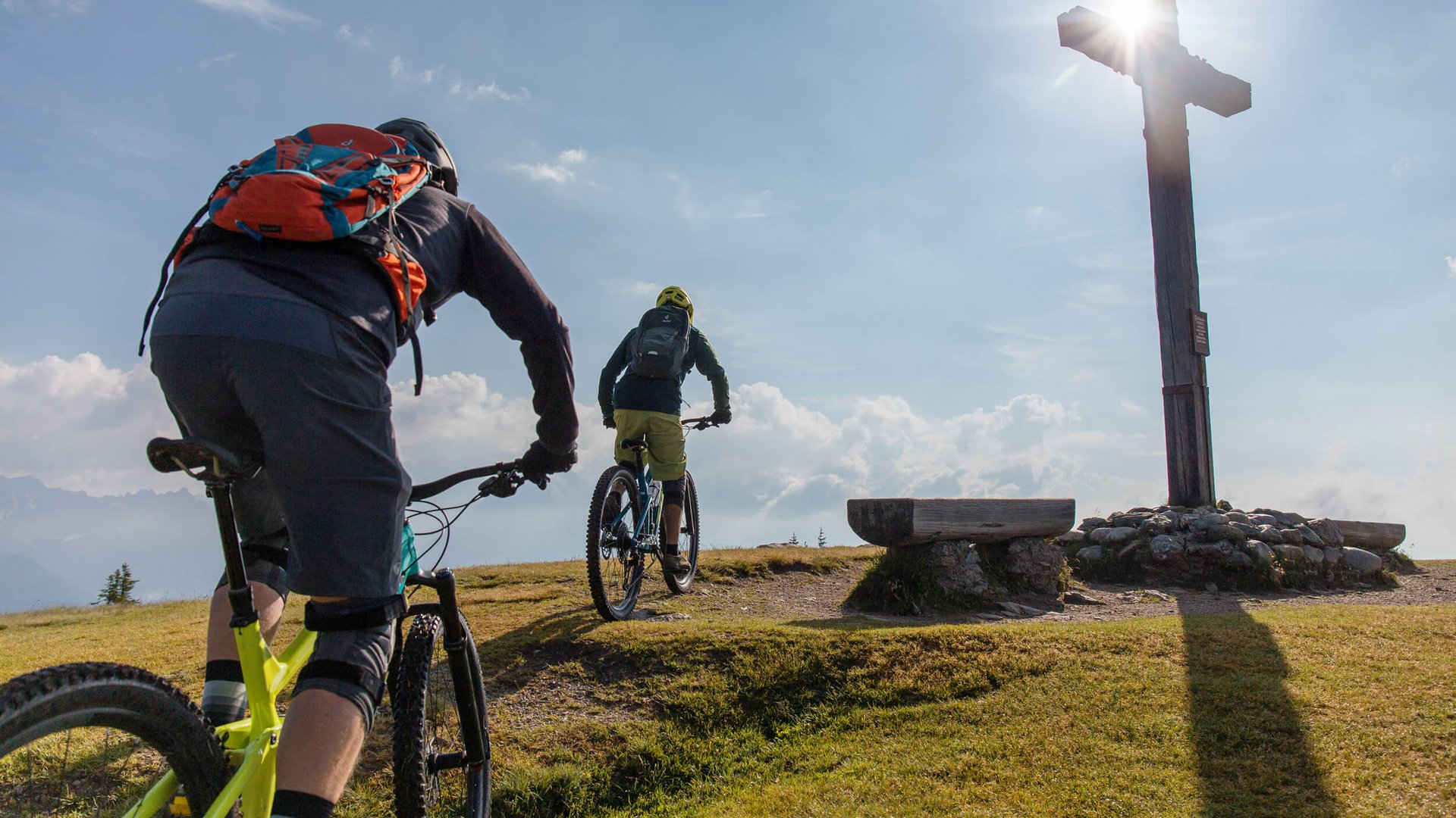 Zwei Mountainbiker fahren zu einem Gipfelkreuz bei Sonnenlicht