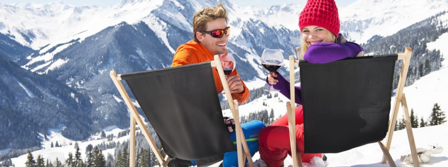 WHITE PEARL MOUNTAIN DAYS Couple sits on deck chairs in snow drinking wine with snowy mountains behind