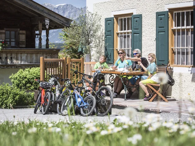 Was steckt hinter dem Umweltengagement von Bikehotels? © Artisual Familie mit Fahrrädern sitzt draußen am Tisch vor einem Haus mit Bergblick