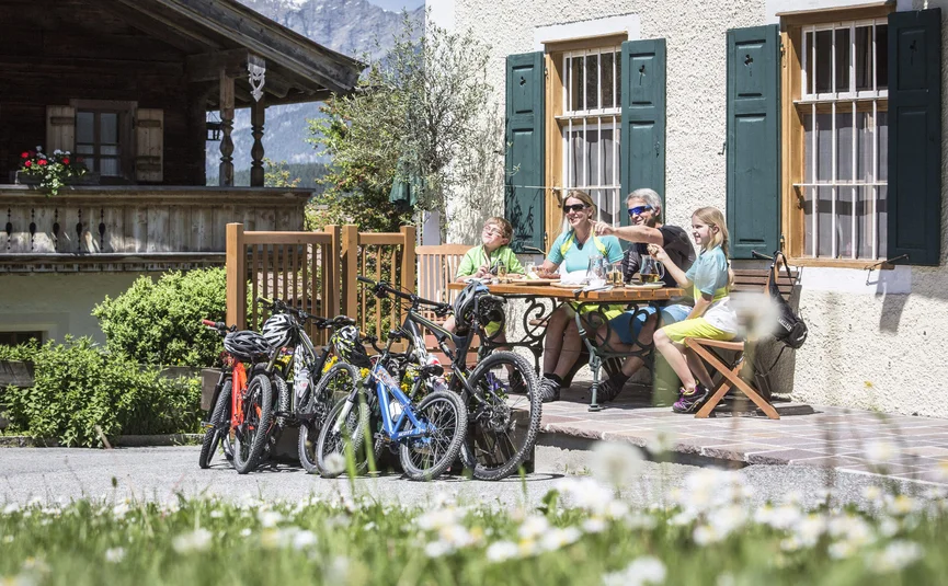 Familie mit Fahrrädern sitzt draußen am Tisch vor einem Haus mit Bergblick