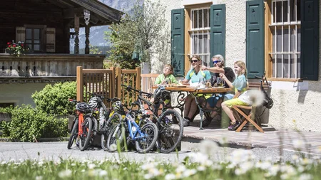 Was steckt hinter dem Umweltengagement von Bikehotels? © Artisual Familie mit Fahrrädern sitzt draußen am Tisch vor einem Haus mit Bergblick