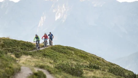 Drei Radfahrer auf Bergweg mit grüner Wiese und Bergen im Hintergrund
