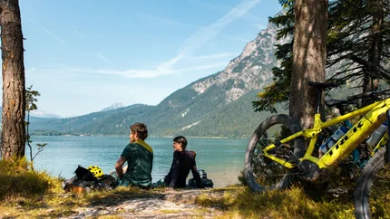 Zwei Radfahrer sitzen am Seeufer vor Berglandschaft
