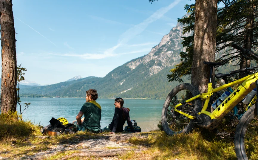 Zwei Radfahrer sitzen am Seeufer vor Berglandschaft