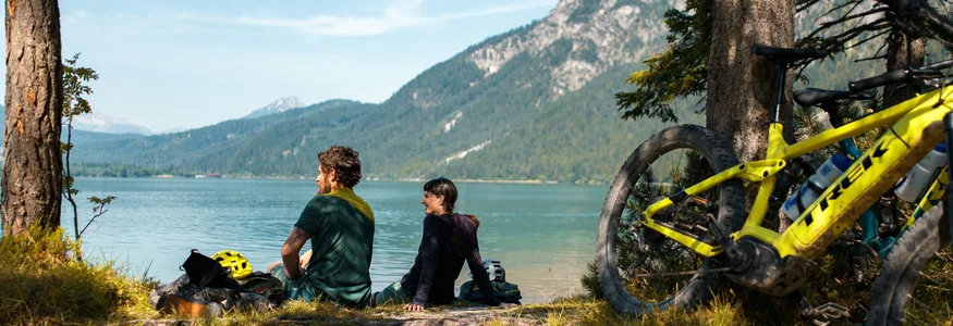 Zwei Radfahrer sitzen am Seeufer vor Berglandschaft