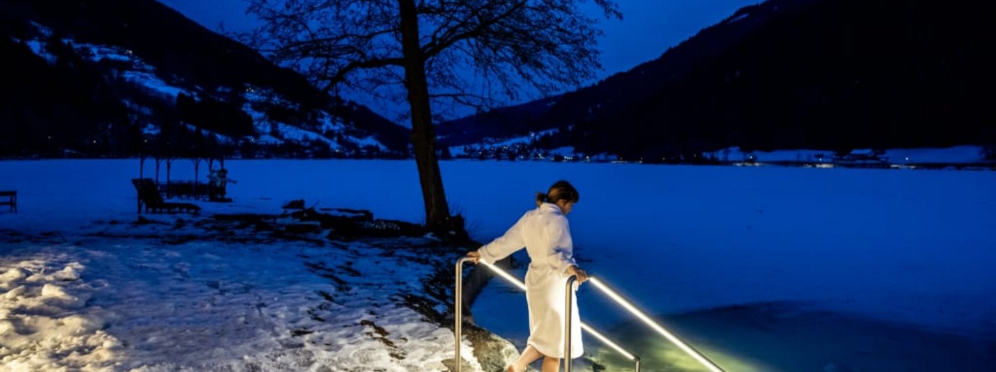 Woman in bathrobe entering icy lake at night with snowy shore