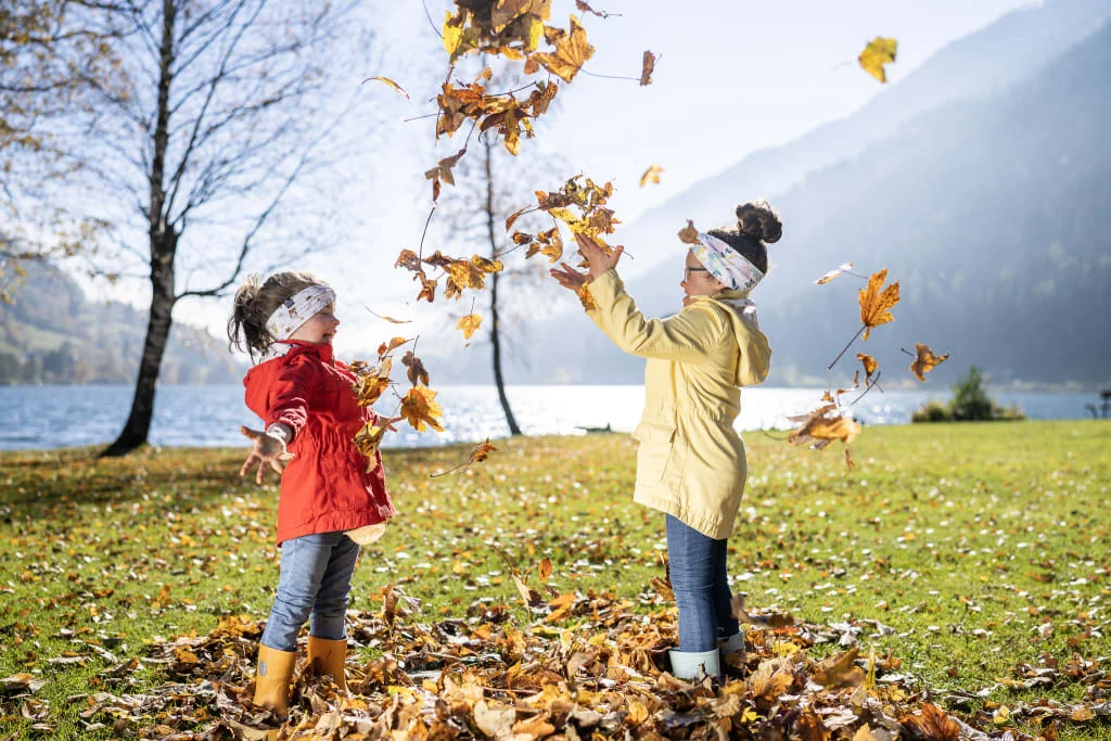 Zwei Kinder spielen mit Herbstblättern am Seeufer bei Sonnenschein