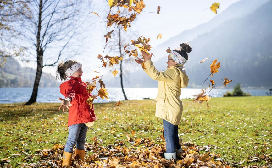 Zwei Kinder spielen mit Herbstblättern am Seeufer bei Sonnenschein