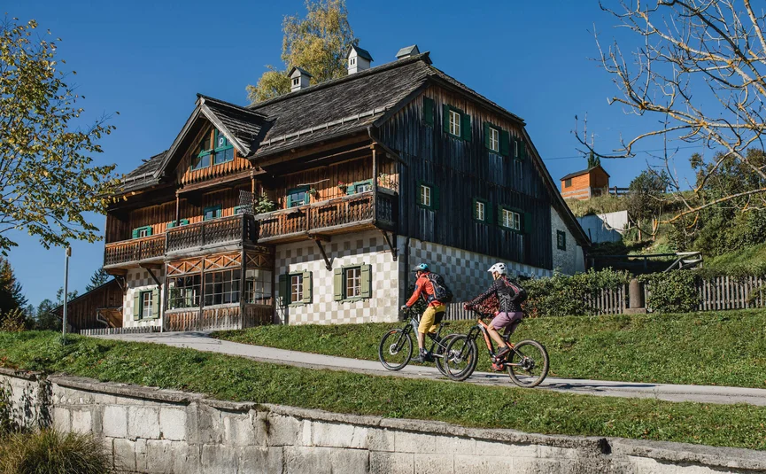 Two cyclists riding past a traditional wooden house under clear sky