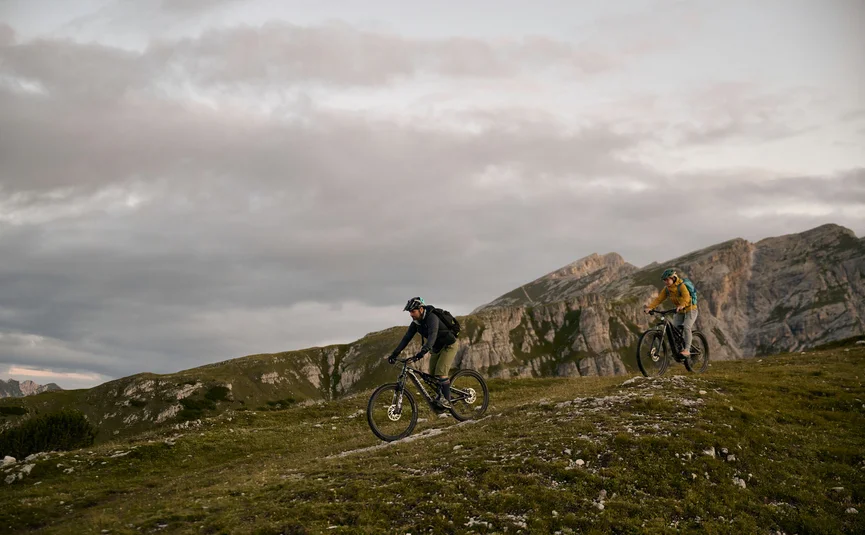 HIRBEN Naturlaub **** © David Rasche Two mountain bikers riding on a grassy hill with mountains in the background