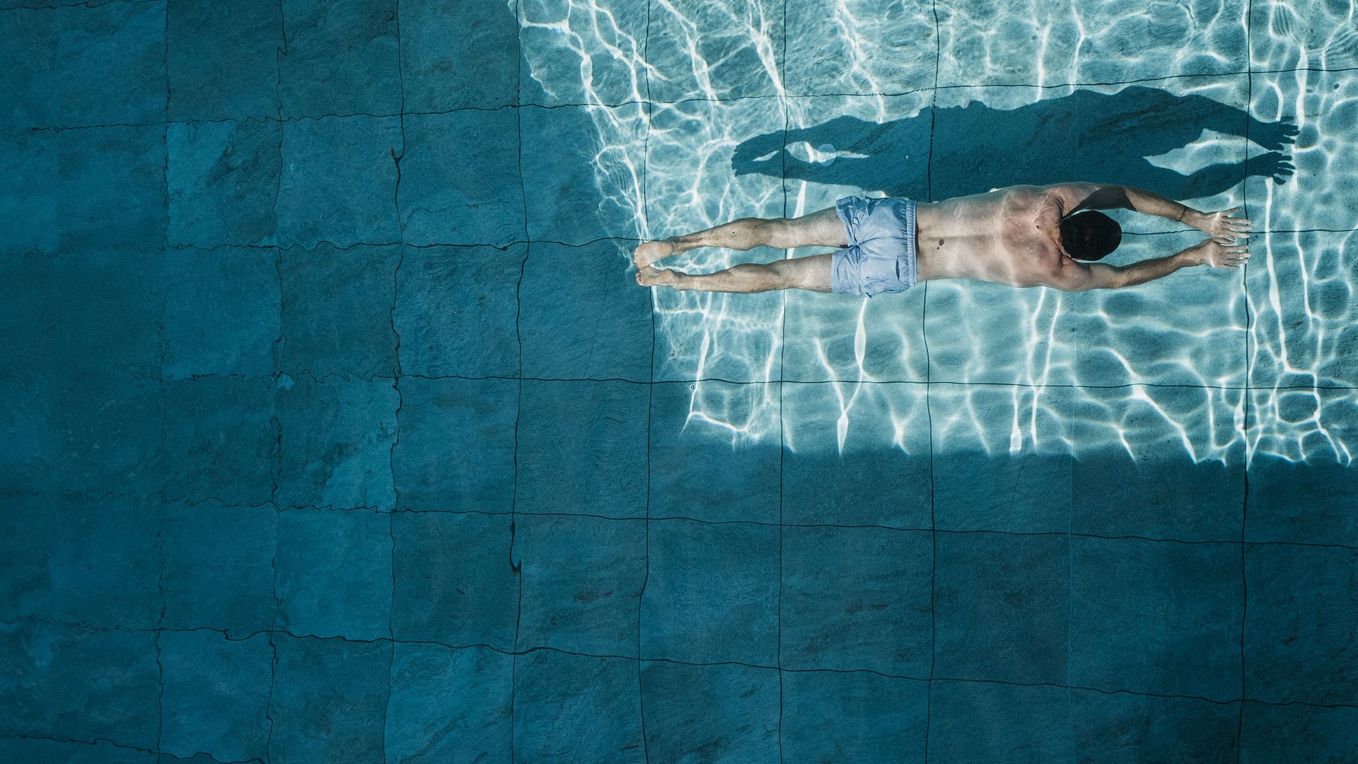 Mann schwimmt auf dem Bauch in klarem Wasser eines Pools mit Kacheln