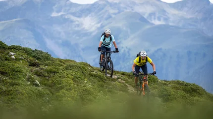 Two mountain bikers riding on a trail with alpine mountains in the background