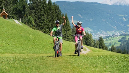 Zwei Radfahrer fahren auf einem grasbewachsenen Bergweg mit erhobenen Armen