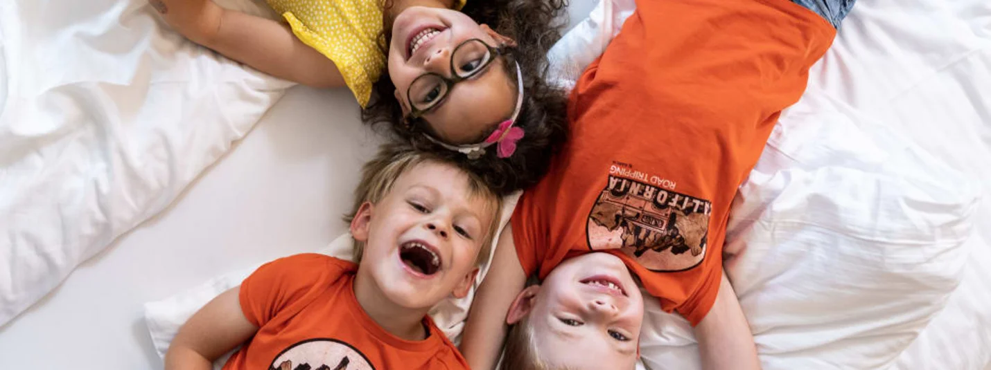 Drie lachende kinderen liggend op een bed met wit beddengoed