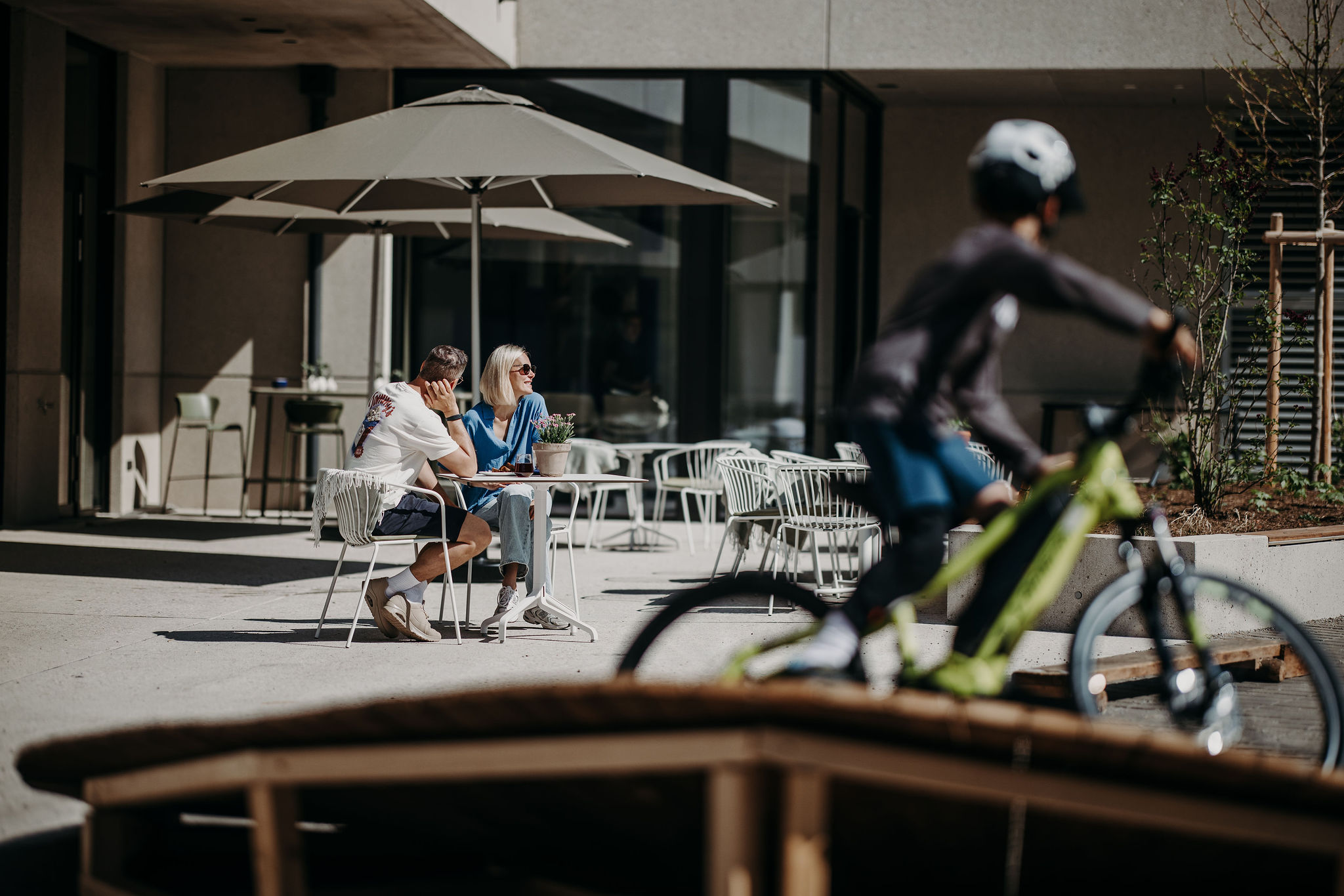 Koppel zit buiten aan tafel onder parasol, fietser rijdt voorbij