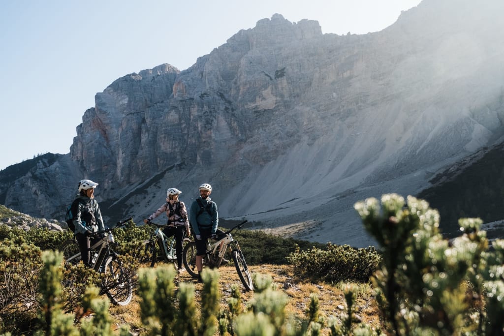 Three cyclists standing with mountains in the background on a trail