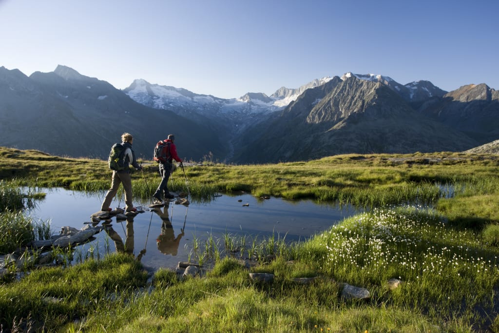 Zwei Wanderer überqueren einen kleinen Bach in einer bergigen Landschaft