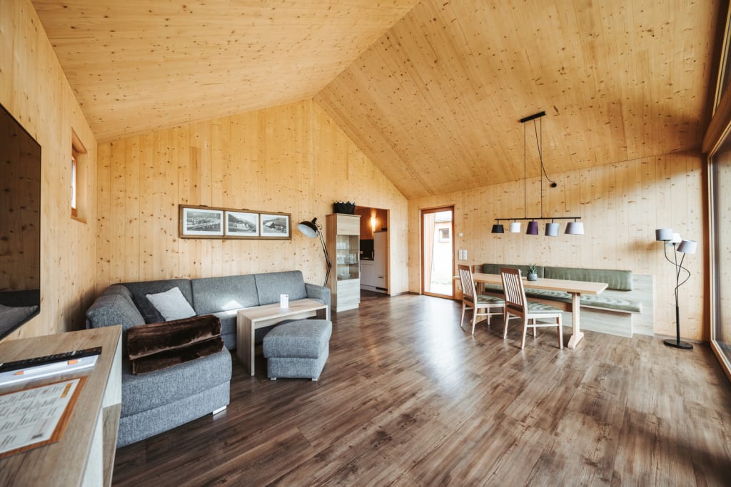Chalet-only Herbst Gemütliches Wohnzimmer mit Holzwänden, grauem Sofa und Essbereich mit Tisch und Stühlen
