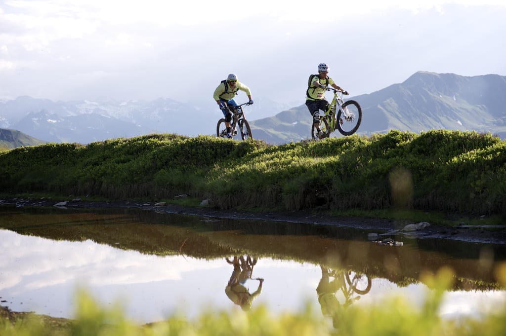 BIKE BREAK (7 Nächte) Zwei Mountainbiker fahren auf einem Hügel vor Bergkulisse mit Wasserreflexion