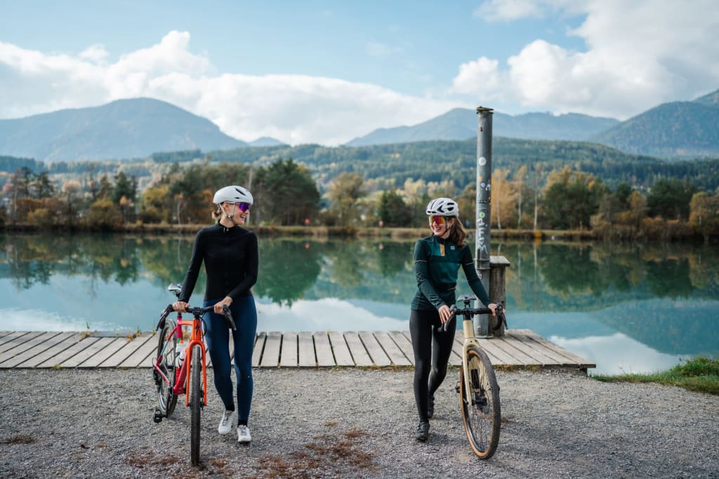 Two female cyclists with bikes by lake shore with mountains in background