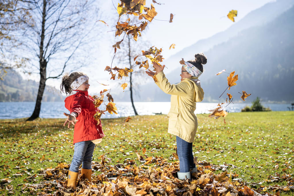 Brennseehof Happy Family Woche Zwei Kinder spielen mit Herbstblättern am Seeufer bei Sonnenschein