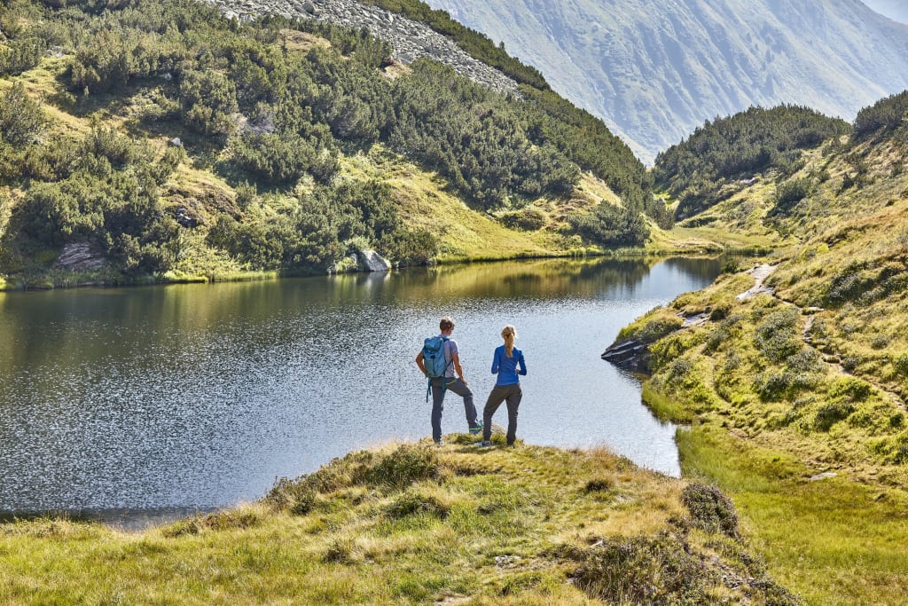 Lamm-Aktiv-Wochen Zwei Wanderer stehen am Ufer eines Bergsees in einer grünen Landschaft
