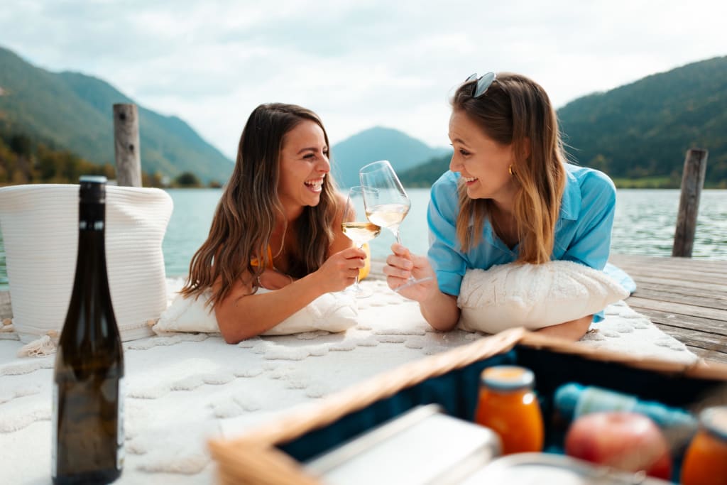 Mädelsauszeit Frühsommer Zwei Frauen stoßen mit Weißweingläsern an am See mit Picknickkorb