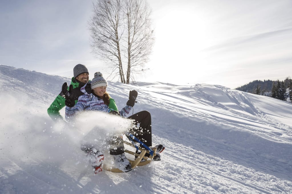 Zwei Personen rodeln den verschneiten Hang hinunter bei sonnigem Wetter