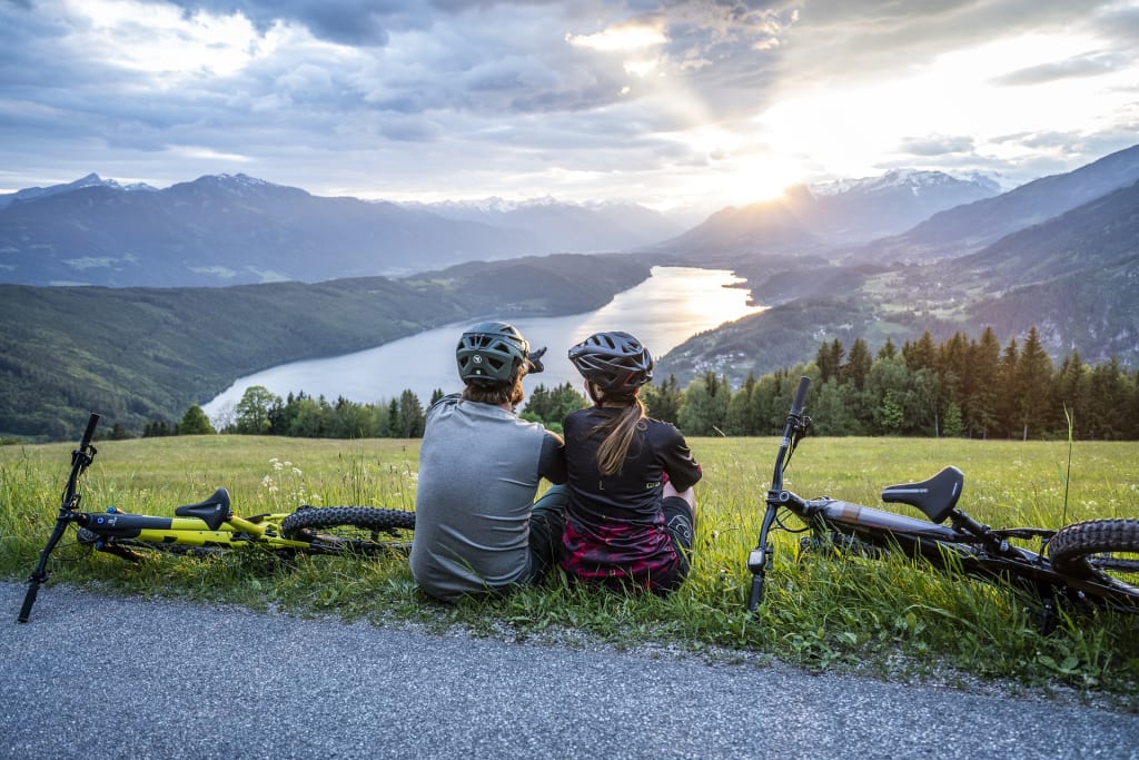 NockBike mit Genuss Twee fietsers zitten op gras en kijken naar een meer en bergen bij zonsondergang