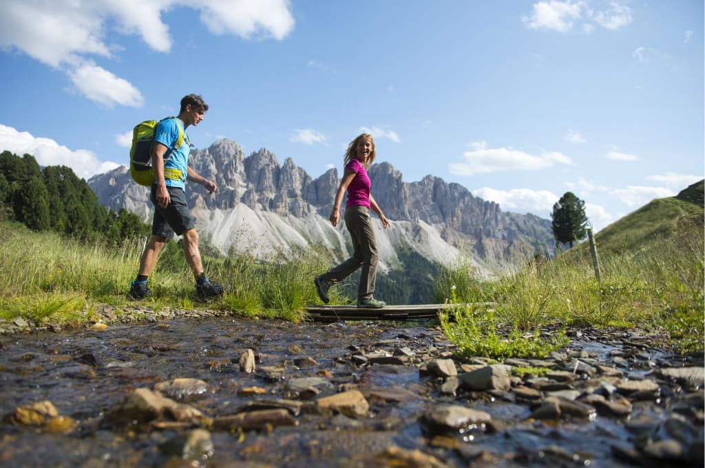 Zwei Menschen wandern über eine Holzbrücke an einem Bergbach mit Alpen im Hintergrund