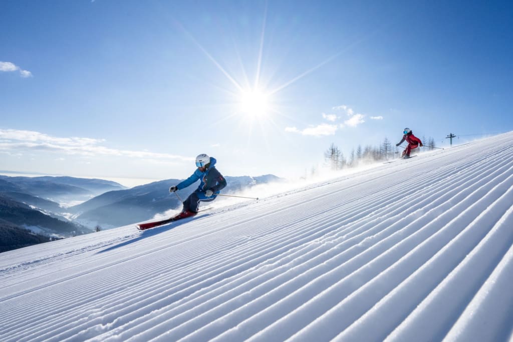 Zwei Skifahrer fahren bei Sonnenschein den frisch präparierten Hang hinunter