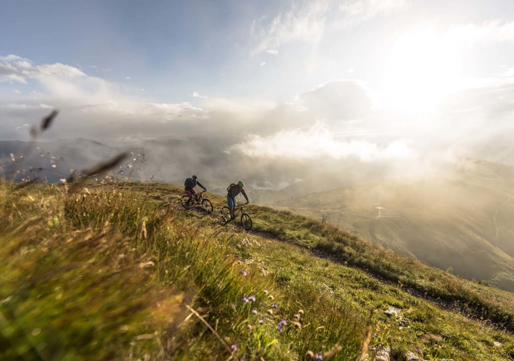 Two mountain bikers riding on a sunny mountain trail with fog in the background