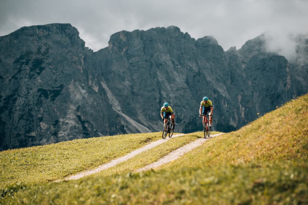 Two cyclists riding on a mountain trail with rocky cliffs and cloudy sky