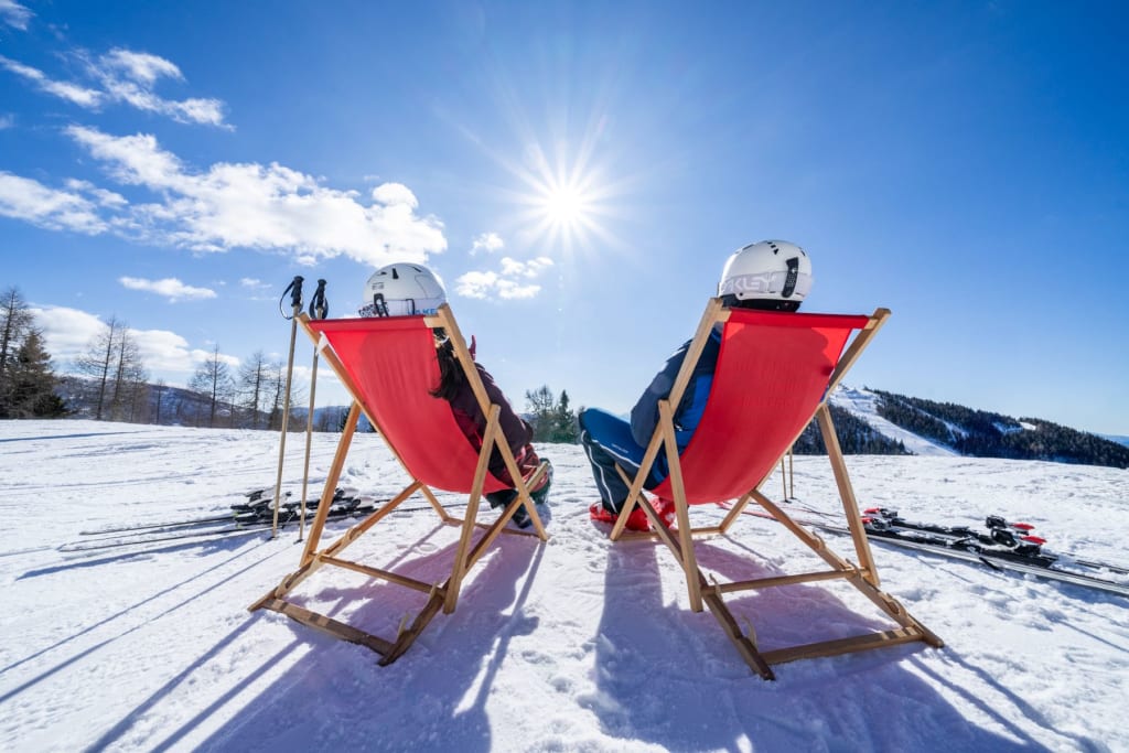 Zwei Skifahrer sitzen in roten Liegestühlen in schneebedeckter Landschaft bei Sonnenschein