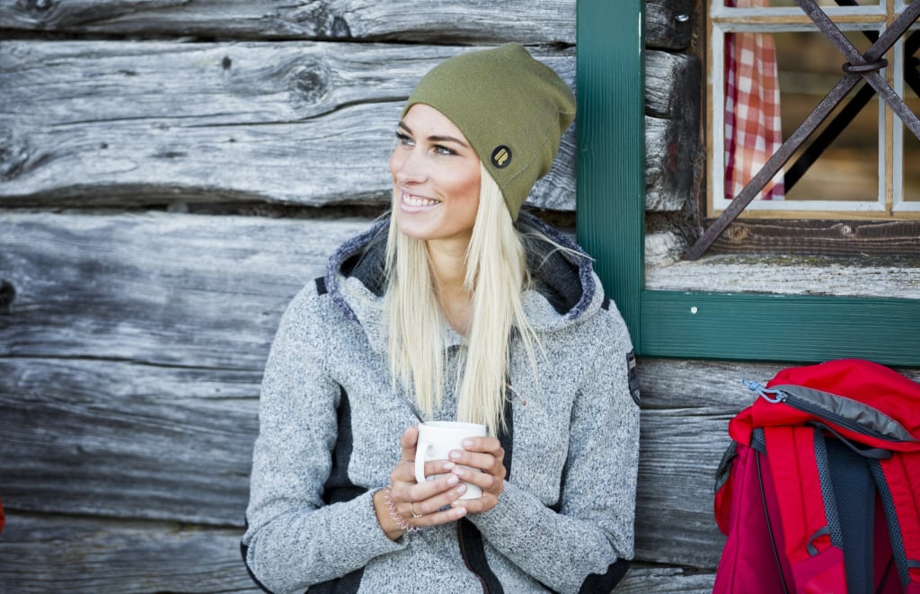 Frau mit grüner Mütze und grauem Pullover trinkt Kaffee draußen am Holzhäusern
