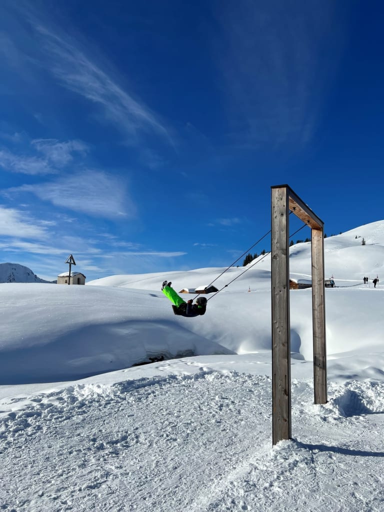 Person schaukelt auf Holzschaukel im verschneiten Bergland unter blauem Himmel
