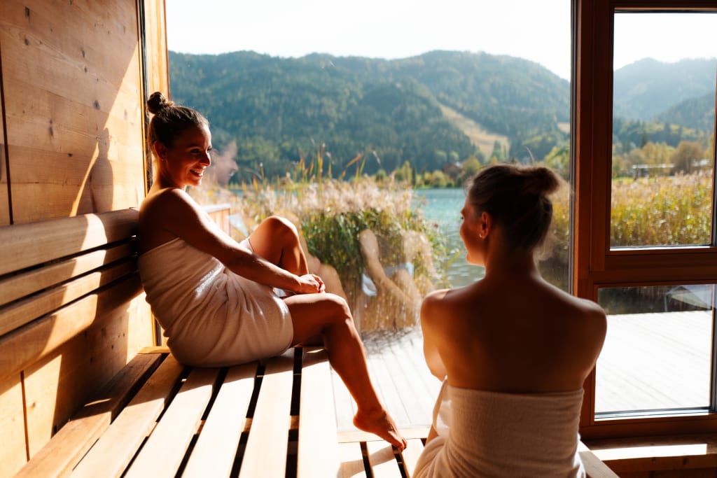 MAI SAUNATAGE Zwei Frauen in Saunatüchern sitzen in einer Sauna mit Seeblick