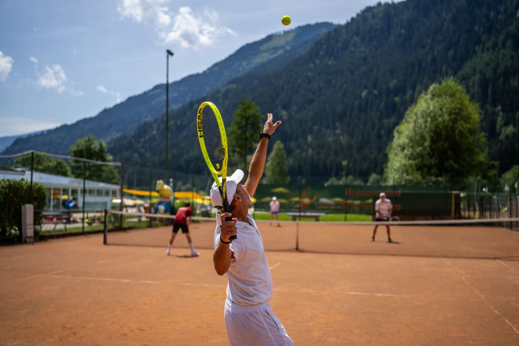 Mann in Weiß serviert Tennisball auf Sandplatz mit Bergen im Hintergrund