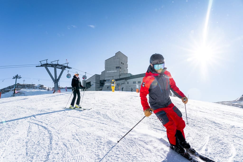 Zwei Skifahrer auf verschneitem Hang mit Seilbahn und Bergstation im Hintergrund