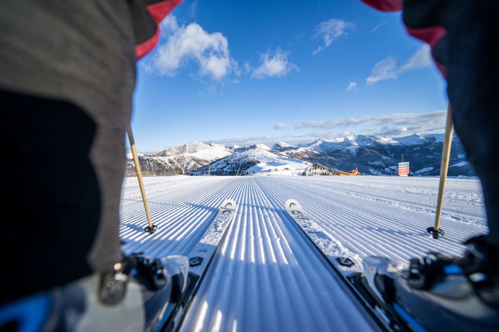 Blick von Skiern auf frisch gespurte Skipiste mit Bergen und blauem Himmel