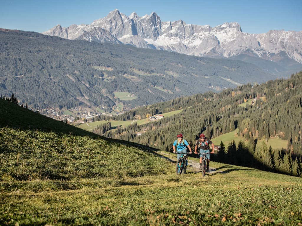 Zwei Mountainbiker fahren auf einem Pfad in den Bergen mit schneebedeckten Gipfeln im Hintergrund