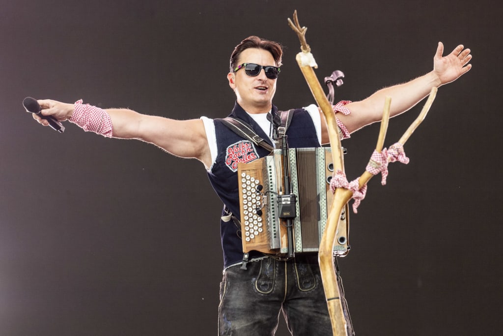 Man on stage with accordion and microphone, arms spread wide